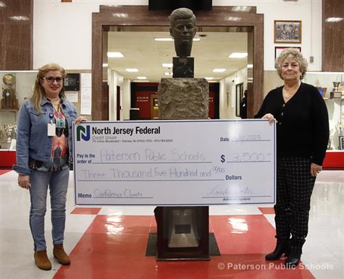 Two women hold a large prop check from North Jersey Federal Credit Union of $3,500 while standing in a large room.